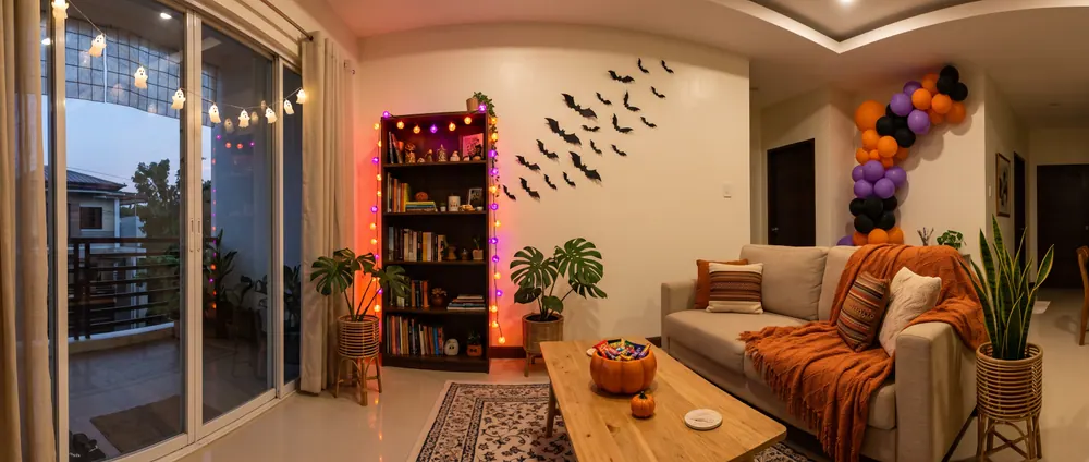 AI sample render of a cozy living room styled for a cute Halloween with orange and purple fairy lights on a bookshelf, glowing ghost lights by the window, black paper bats on the wall, a ceramic pumpkin bowl and a balloon garland