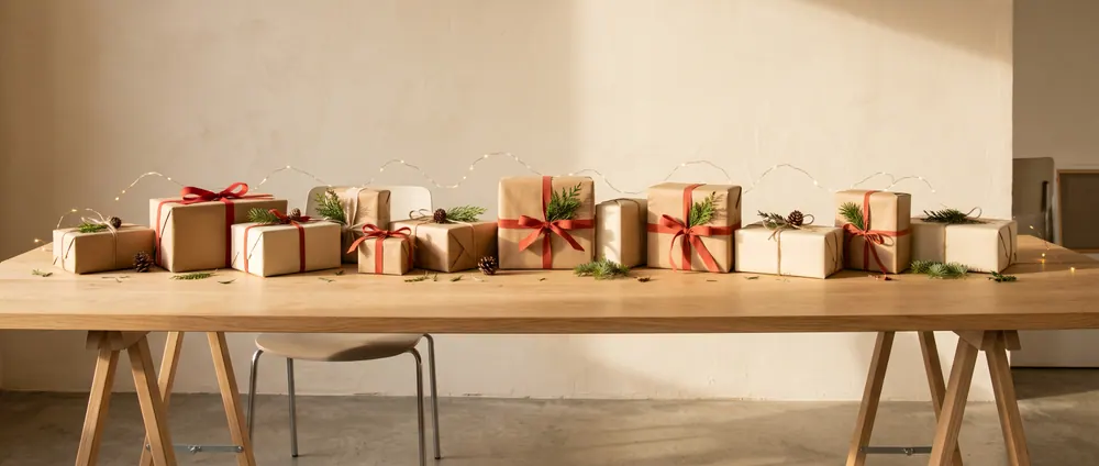 AI sample render of an office gift-exchange table with kraft-wrapped gifts, red and terracotta ribbons, pine sprigs and warm string lights