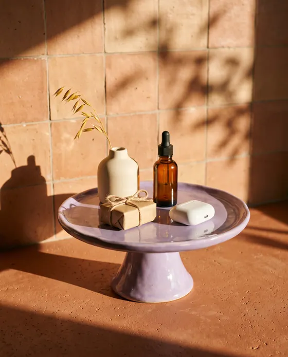 AI sample render of a lilac ceramic stand in warm sunlight holding a small vase, a dropper bottle, wireless earbuds and a wrapped gift against terracotta tiles